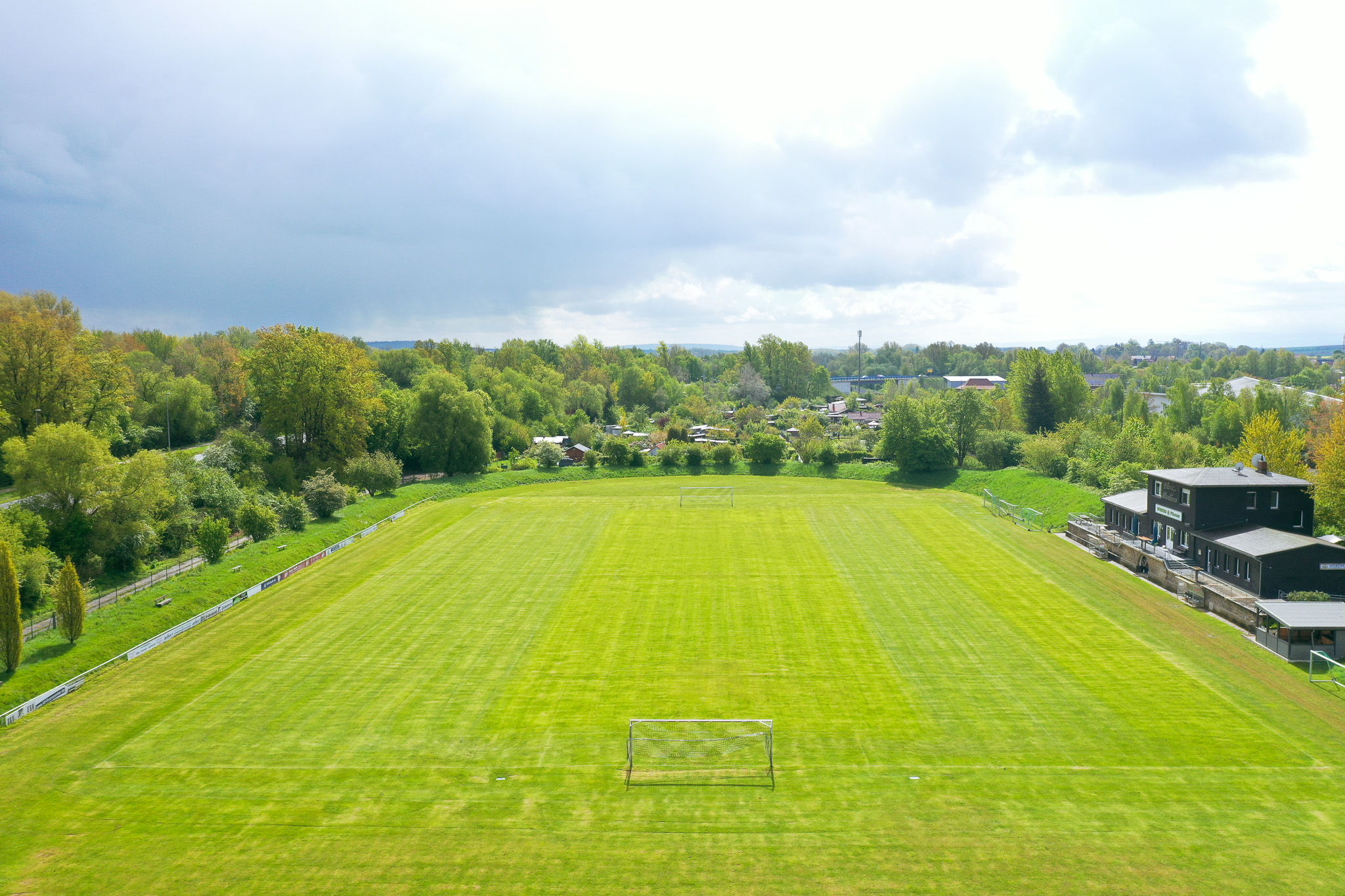 Fußballplatz in Niedersachsen mit frisch gemähtem Rasen, eingerahmt von Bäumen einem modernen Vereinsheim mit Terrasse.