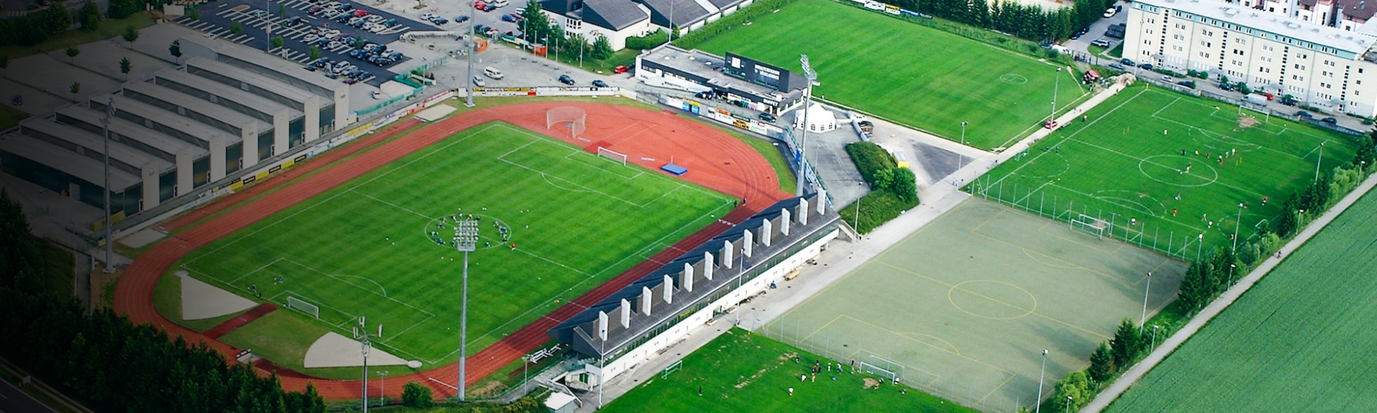 Sportzentrum in Oberösterreich aus der Vogelperspektive mit Stadion, Trainingsplätzen und einer Laufbahn, eingebettet in eine städtische Umgebung mit angrenzenden Feldern und Verkehrsstraßen.
