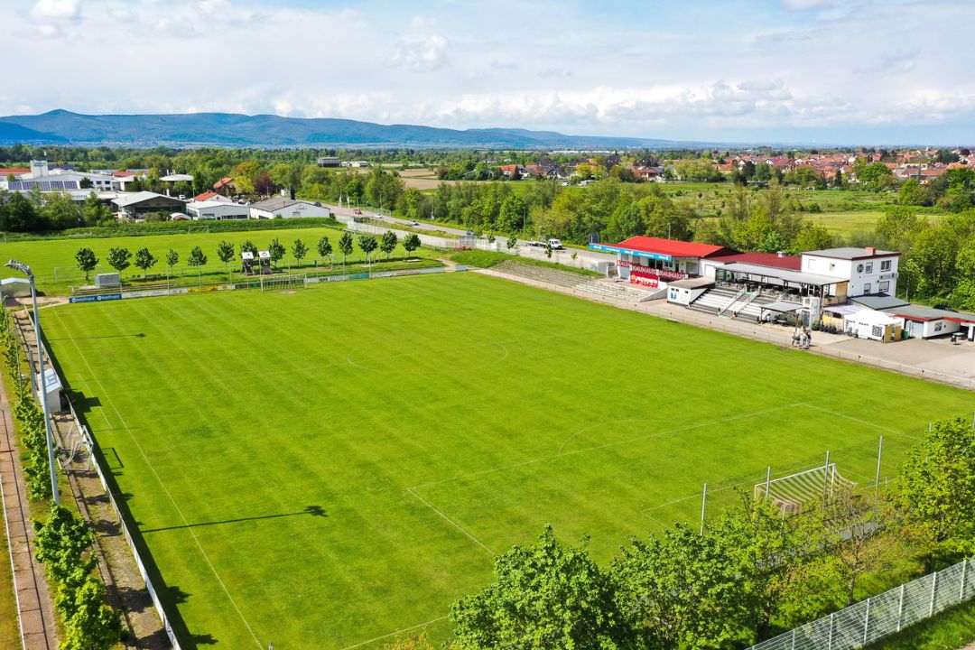 Luftaufnahme eines gepflegten Naturrasen-Fußballplatzes mit angrenzendem Vereinsheim und Tribüne, eingebettet in eine grüne Landschaft mit Blick auf eine Stadt und Hügel im Hintergrund in Rheinland-Pfalz.