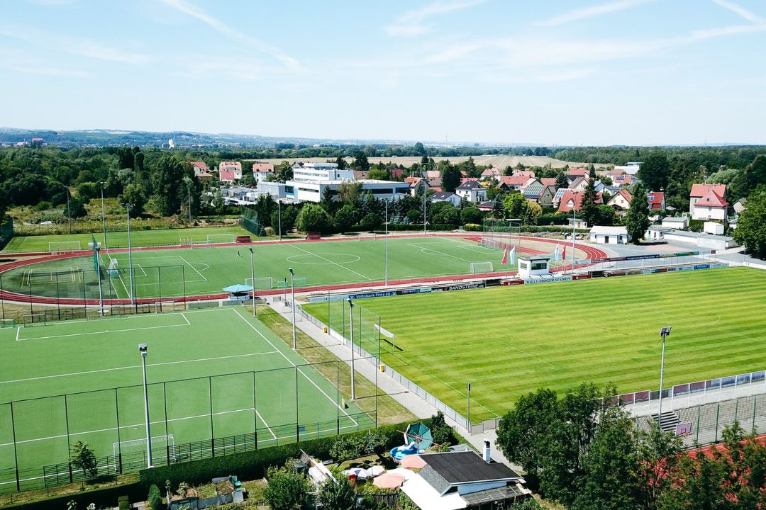Sportanlage mit Fußballplätzen, Laufbahn und Stadion, umgeben von Wohnhäusern und grüner Landschaft.