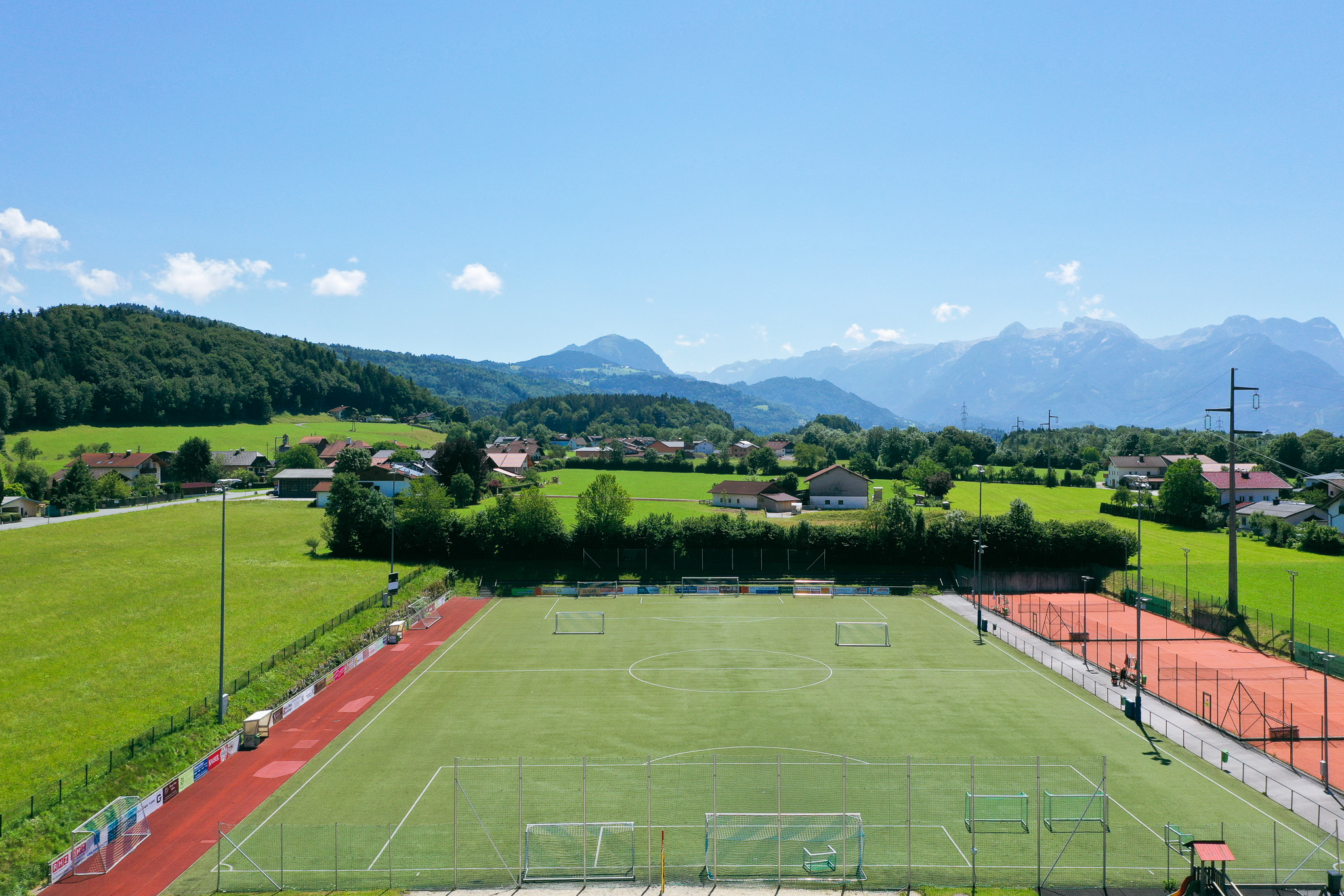 Fußballplatz in Salzburg, umgeben von grünen Wiesen, kleinen Häusern und malerischen Bergen im Hintergrund, unter blauem Himmel.