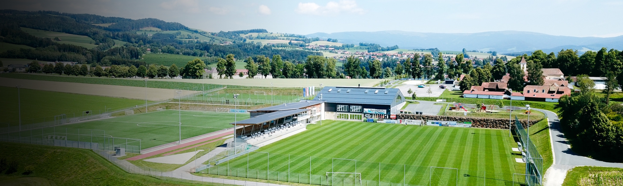 Panoramaaufnahme eines Fußballplatzes in der Steiermark, eingebettet in eine idyllische Hügellandschaft, mit Tribüne, Trainingsplatz und Vereinsgebäude.