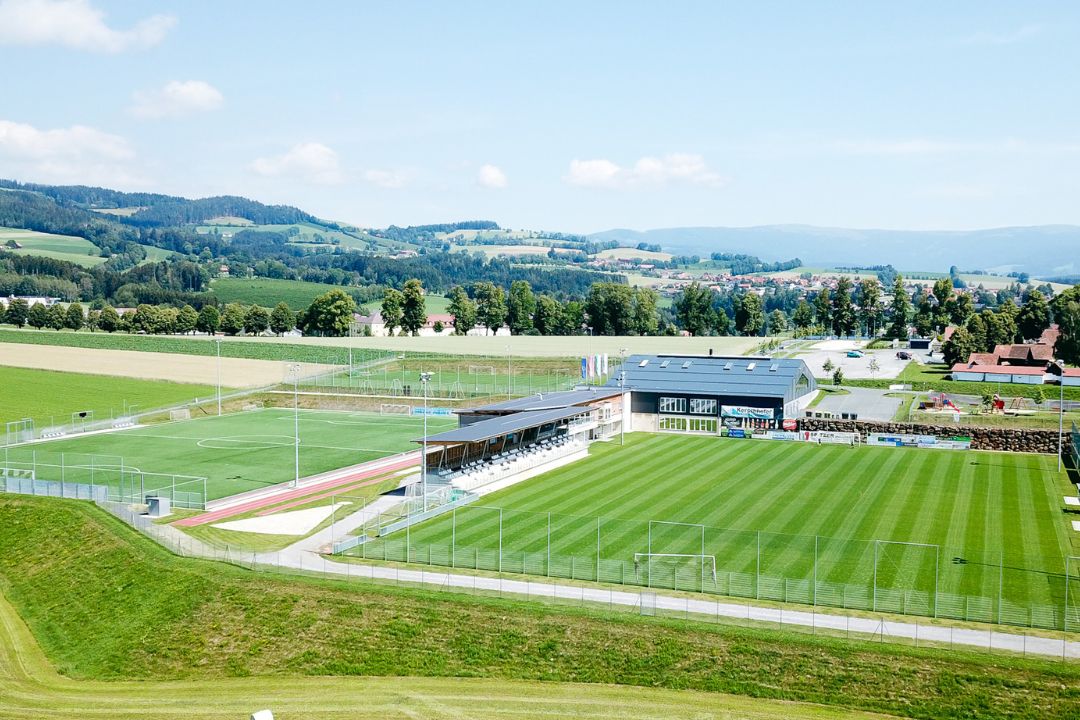 Moderne Fußballanlage in der Steiermark, umgeben von grünen Hügeln und Wiesenlandschaft, mit gepflegtem Naturrasenplatz, Tribüne und Sportgebäude im Hintergrund.