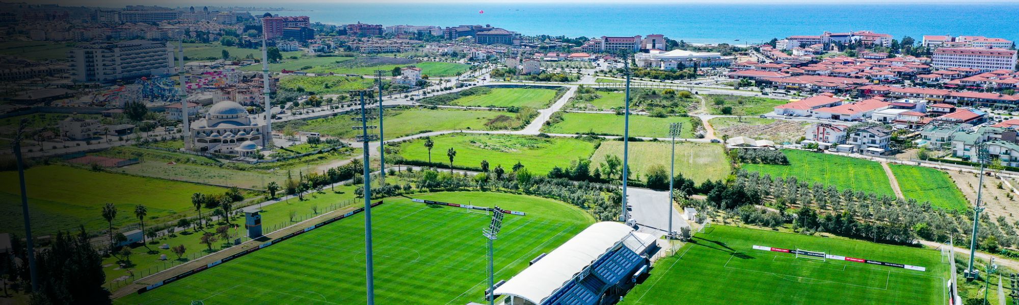 Luftaufnahme eines Fußballtrainingslagers in der Türkei mit zwei Rasenplätzen, Tribüne und Blick auf die Stadt und das Meer.