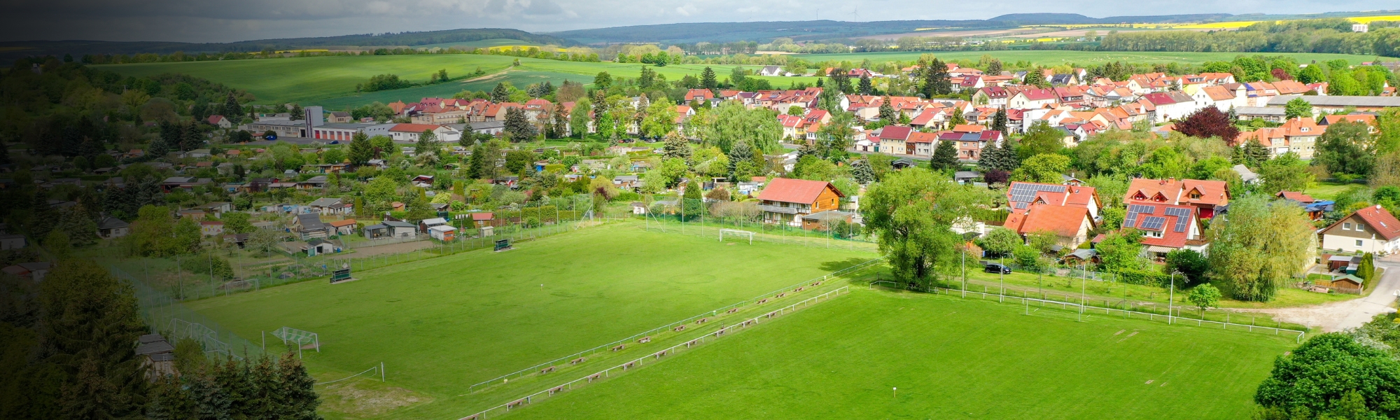 Fußballplatz in ländlicher Umgebung in Thüringen, umgeben von grünen Wiesen, Kleingärten und einer Dorfkulisse mit roten Dächern.