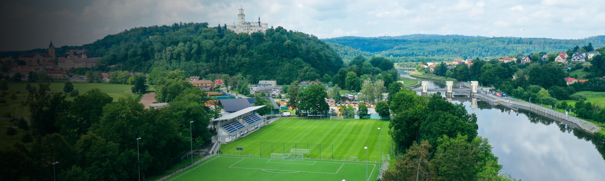 Luftaufnahme eines Fußballplatzes in Tschechien, neben einem Fluss, mit Schloss und Stadt im Hintergrund.