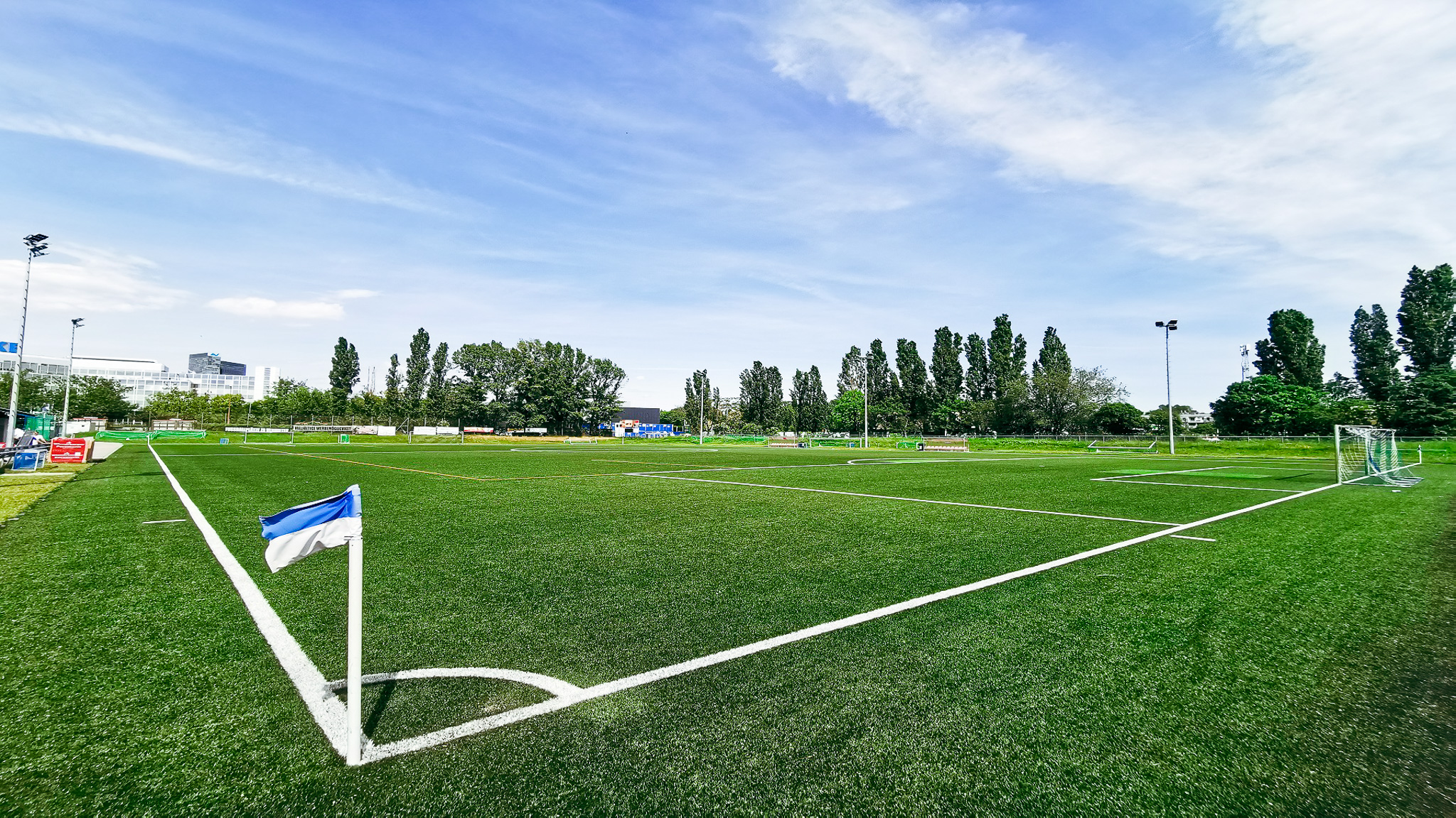 Fußballplatz mit Kunstrasen und blauer Eckfahne in Wien, umgeben von Bäumen und modernen Gebäuden im Hintergrund.