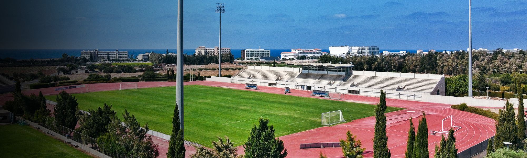 Panoramaaufnahme des Trainingsstadions auf Zypern mit Meerblick, Tribünen und umliegenden Hotelanlagen.
