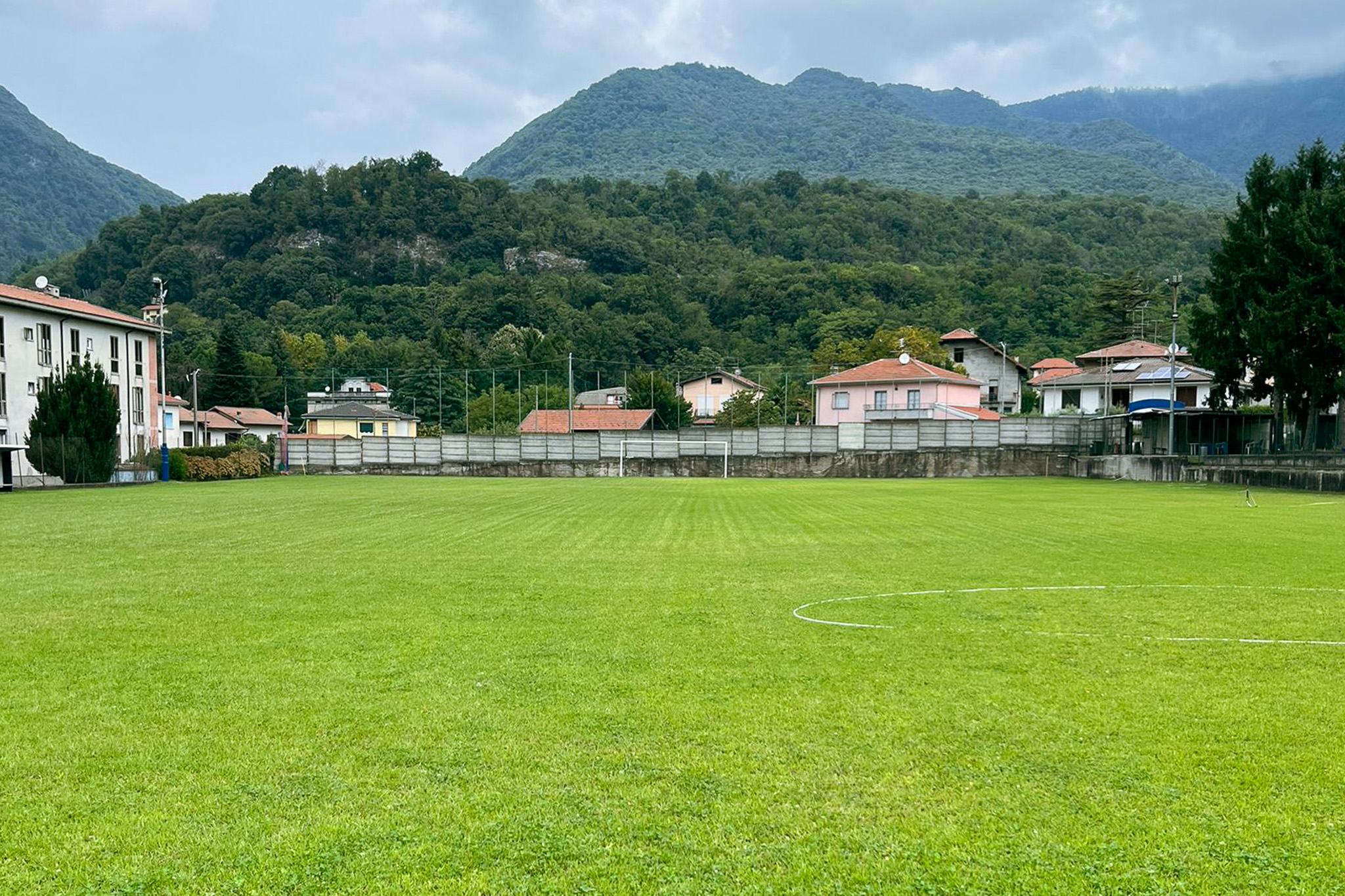 Fußballtrainingsplatz auf Sardinien mit grünem Kunstrasen, Tor, Bänken und Flutlichtmasten, umgeben von Netzen und Berglandschaft.